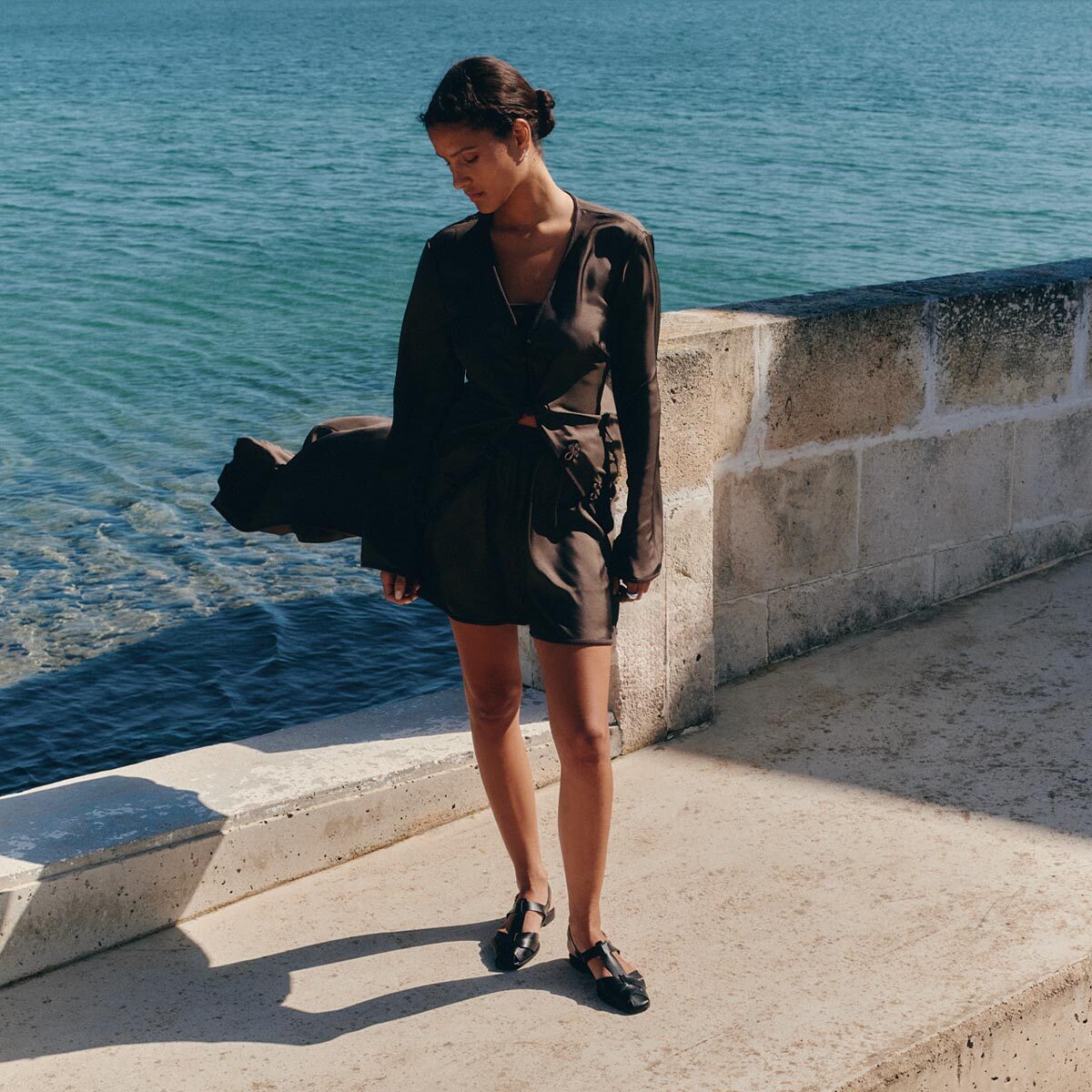 A woman wearing Margaux's The Calypso Fisherman - Black Calf stands on a stone walkway by the serene blue sea, looking down as sunlight casts gentle shadows around her.