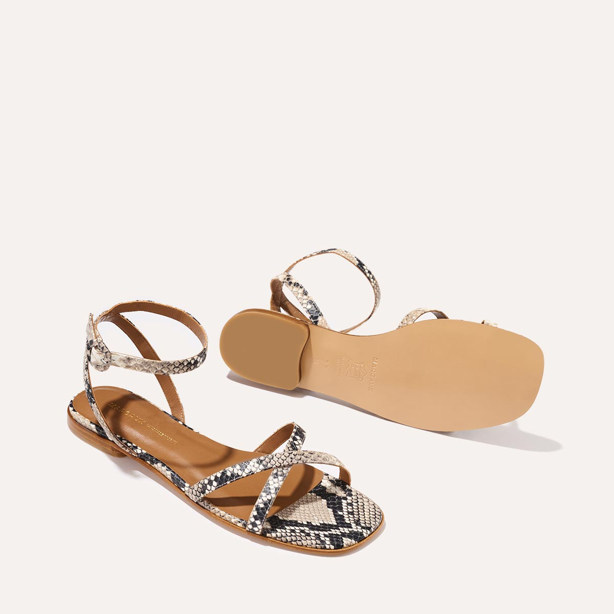 The Margaux Flat Sandal in Natural Python Embossed features a brown sole and slender, snakeskin-patterned straps with an ankle strap—one sandal stands upright while the other reveals its sole, showcasing a barely-there, timeless design.