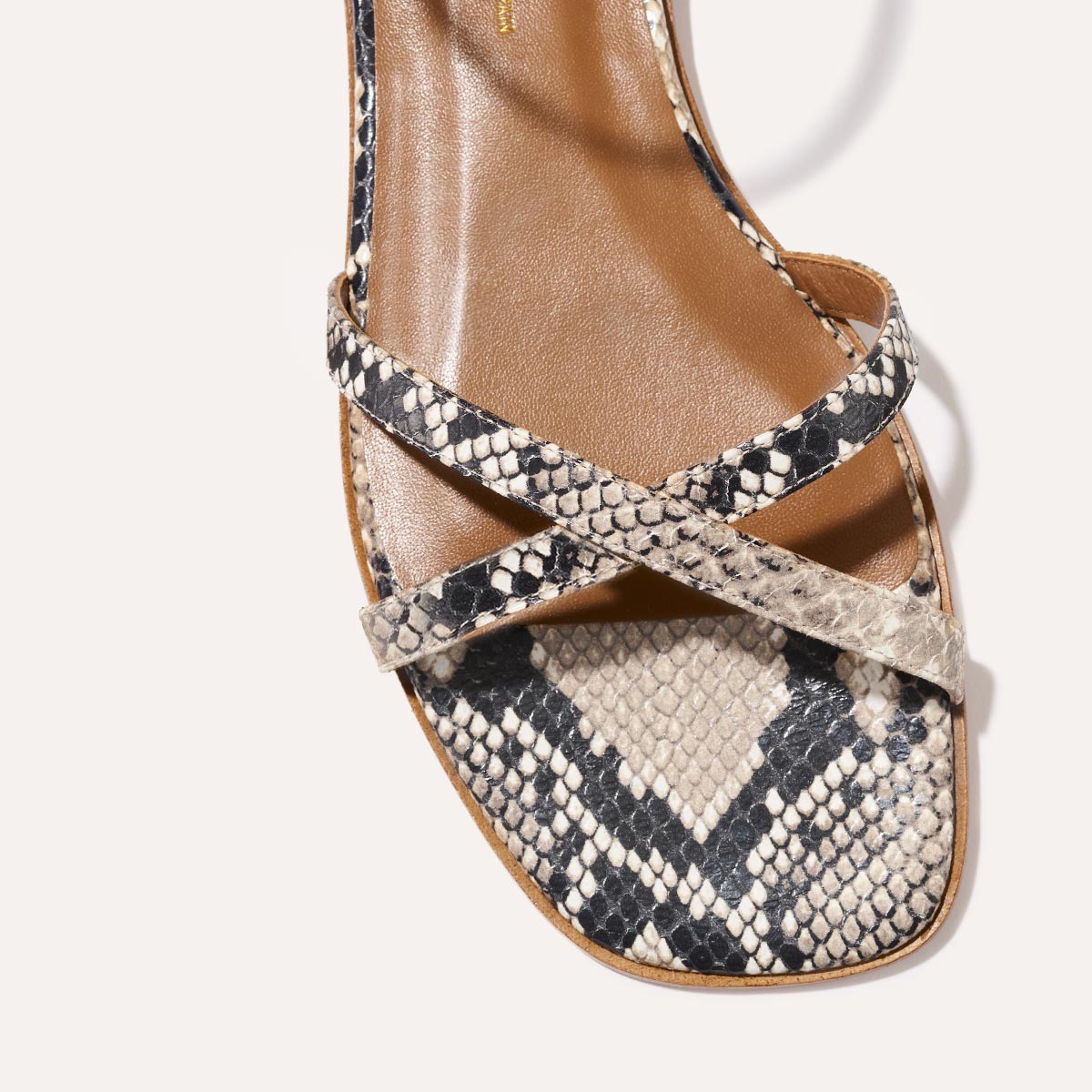 Close-up of The Flat Sandal - Natural Python Embossed by Margaux, featuring a brown sole, criss-cross straps in black, white, and beige python-embossed leather, a foam-cushioned insole, and a square toe for timeless style.