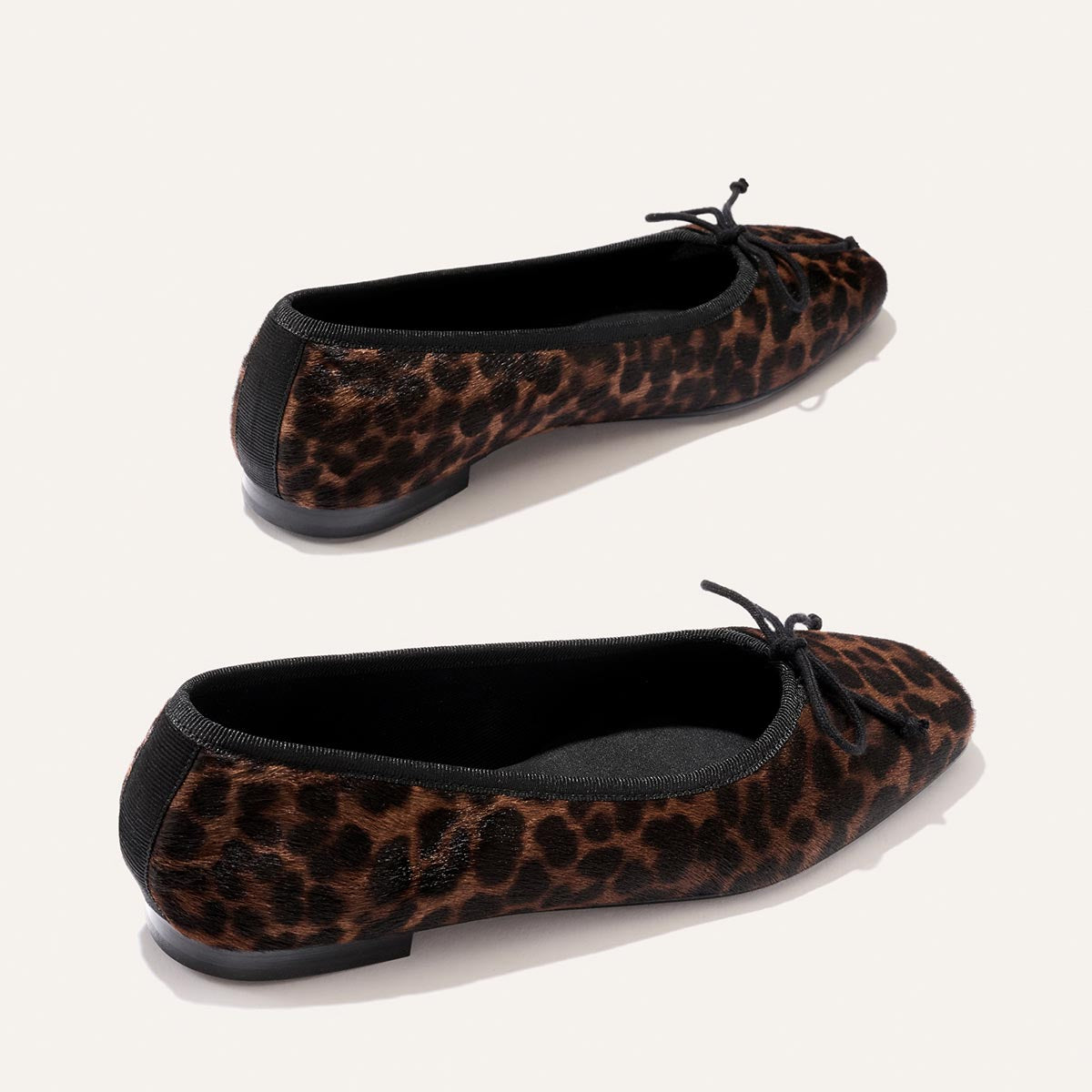 Two pairs of The Fonteyn - Chocolate Leopard Haircalf ballet flats by Margaux, featuring brown and black leopard-print uppers in luxurious haircalf, are handcrafted with a comfort-first design. Each shoe is finished with black trim and a delicate bow at the toe, set against a simple, light background.