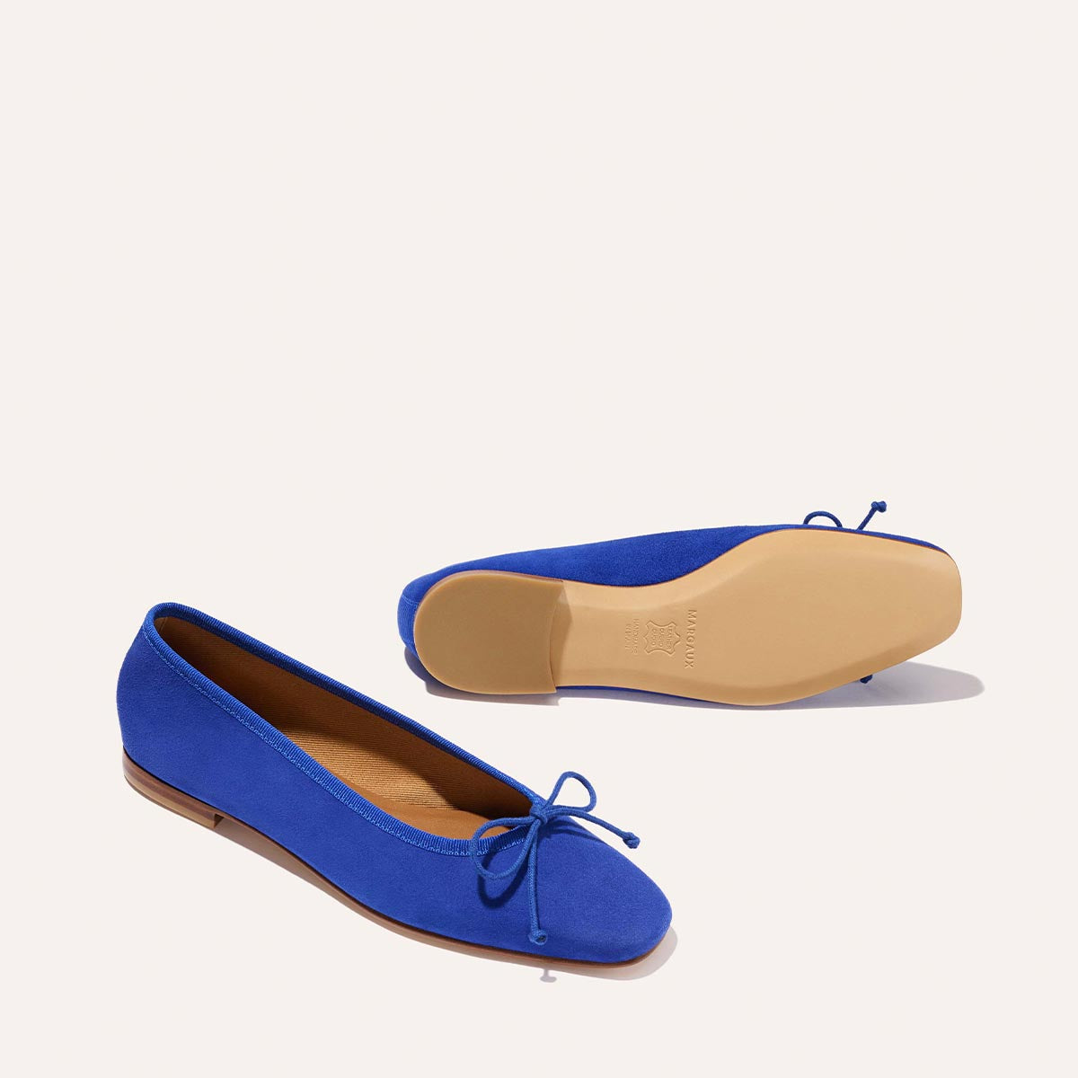 The Fonteyn - Cobalt Suede ballet flats by Margaux feature cobalt blue suede, comfort-first design, and bow details; one shoe stands upright while the other rests on its side to display the beige sole against an off-white background.