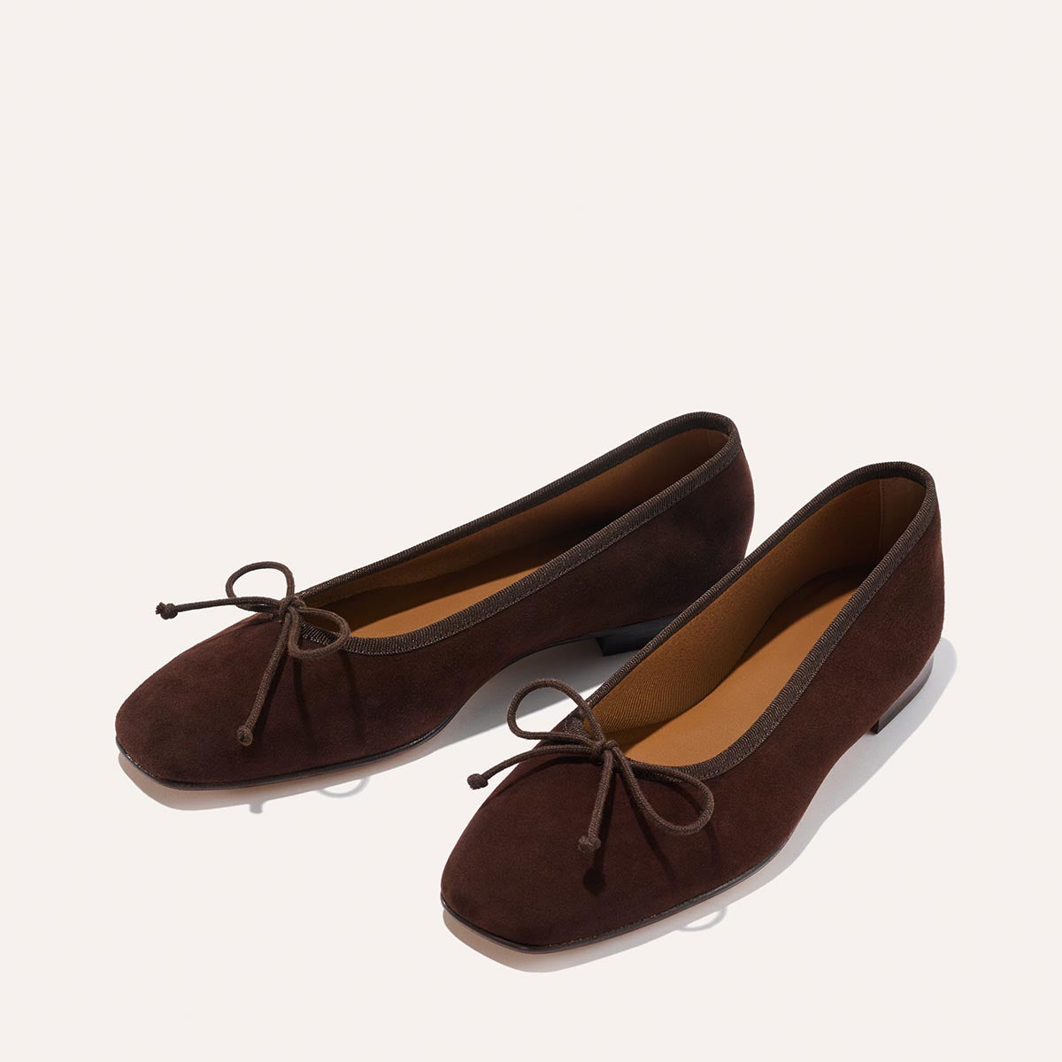 A pair of Margaux's The Fonteyn - Espresso Suede ballet flats, handcrafted in dark brown suede with rounded toes and bow accents, are displayed on a plain light background for timeless comfort.