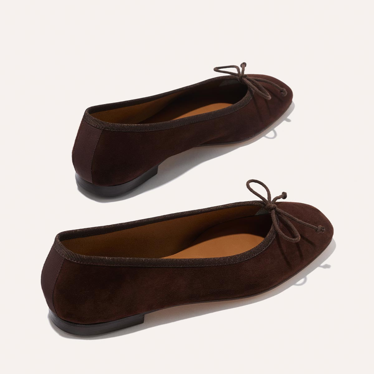The Fonteyn - Espresso Suede by Margaux are dark brown suede ballet flats with bow details and almond-shaped toes, designed for daily comfort. Handcrafted with flat soles, they're shown from a slightly elevated angle on a light background.