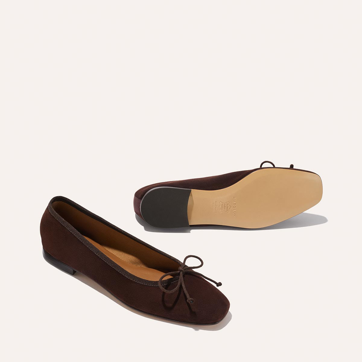 The Fonteyn - Espresso Suede by Margaux: dark brown suede ballet flats with petite front bows, designed for comfort. One shoe stands upright; the other displays its beige sole and low heel against a simple light backdrop.