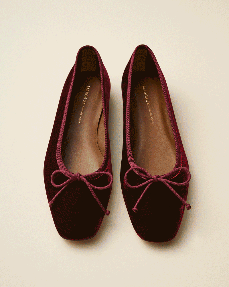 A pair of burgundy velvet ballet flats with square toes and matching bow accents on the front, displayed on a neutral background.