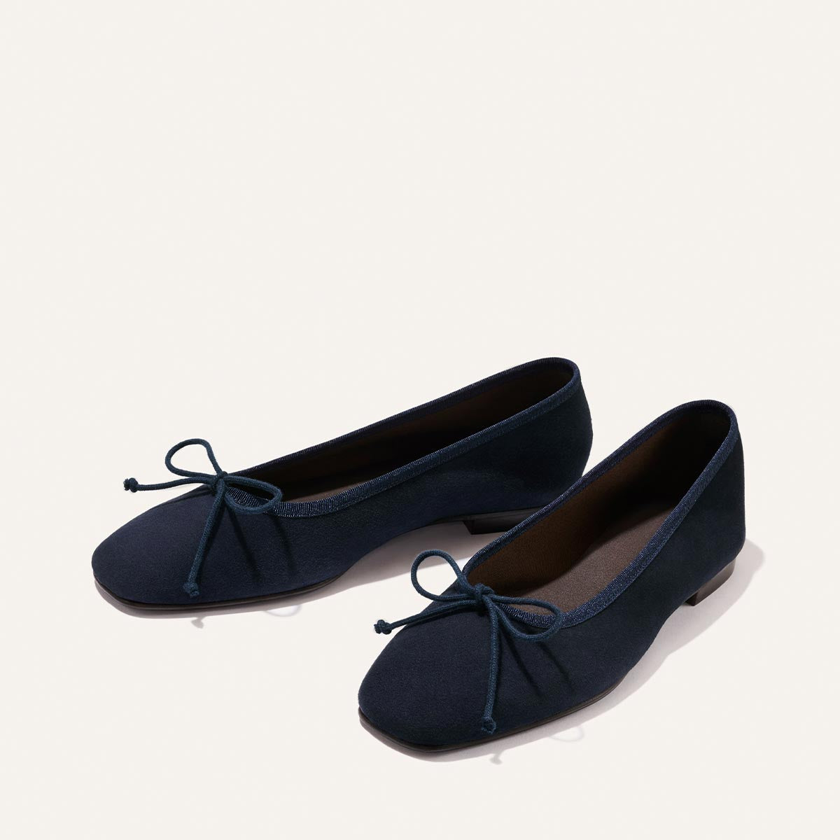The Fonteyn - Indigo Suede by Margaux: a pair of navy blue ballet flats with small bows on the toes, displayed on a light background.