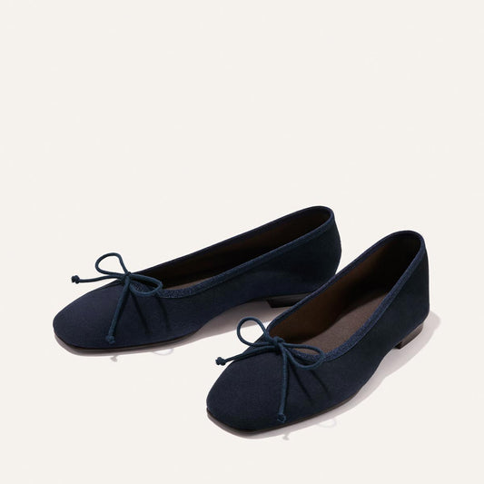The Fonteyn - Indigo Suede by Margaux: a pair of navy blue ballet flats with small bows on the toes, displayed on a light background.