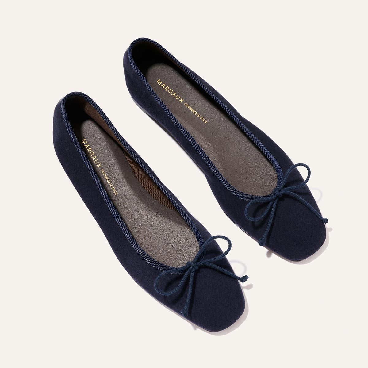 A pair of Margaux's The Fonteyn - Indigo Suede ballet flats with rounded toes and small front bows, shown on a light background. The light brown insoles display the Margaux brand name in gold lettering.