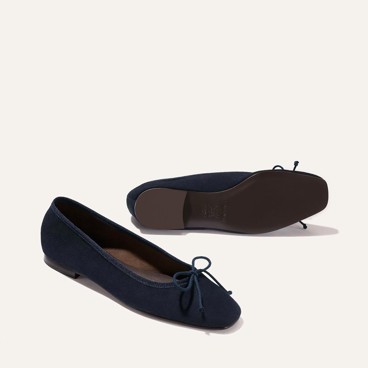 The Fonteyn - Indigo Suede by Margaux: a pair of navy blue ballet flats with a bow on the front; one shoe is upright, the other displays the sole.