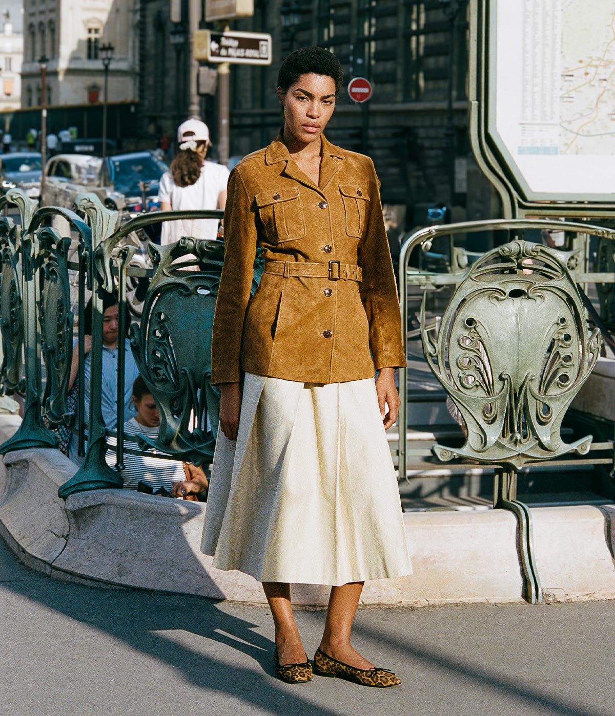 A woman wearing a brown belted jacket, cream midi skirt, and leopard print flats stands near an ornate subway entrance in a city, with people and cars in the background.