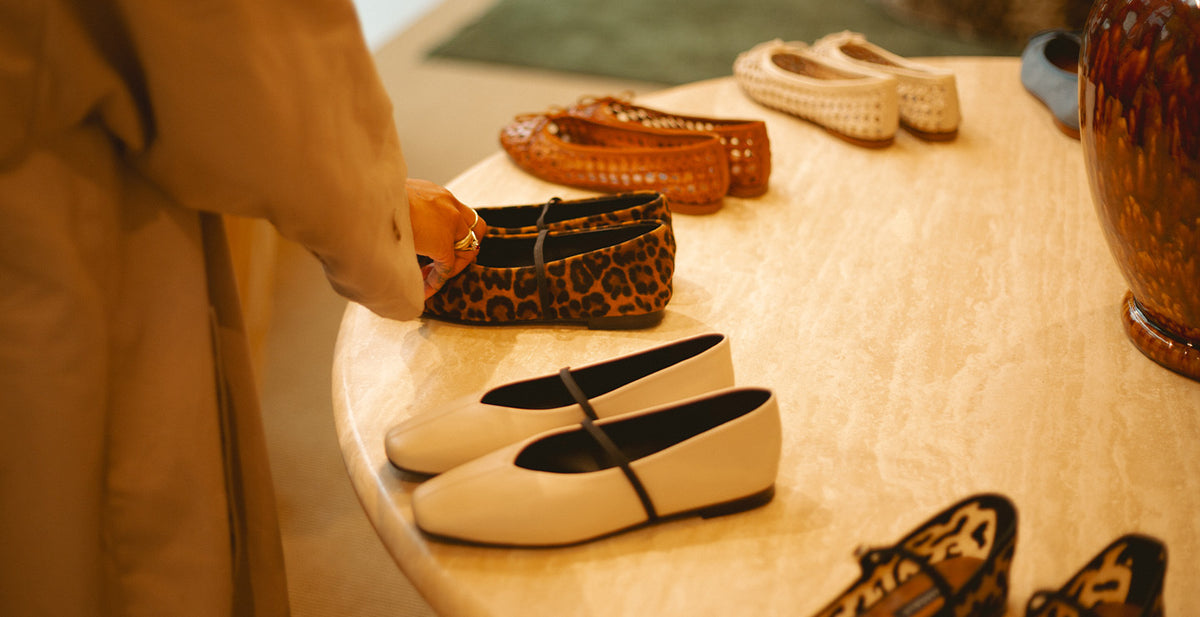 A person reaches for a pair of leopard print flats on a round beige table displaying various styles of womens shoes, including white flats and woven loafers.
