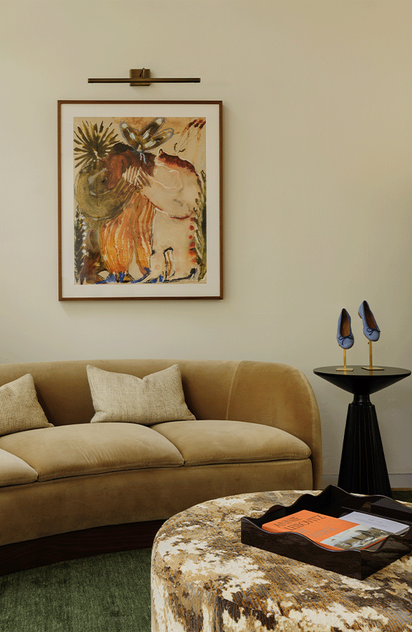 A beige curved sofa with two cushions sits below an abstract framed painting on a cream wall. A round ottoman with a tray and a side table with blue ornaments complete the cozy, modern living room.