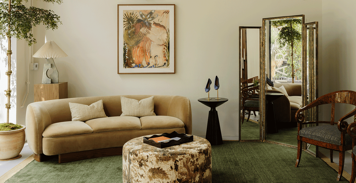 A stylish living room with a beige curved sofa, green rug, abstract art on the wall, round ottoman, side tables, a decorative lamp, potted plants, and a large mirror reflecting natural light.