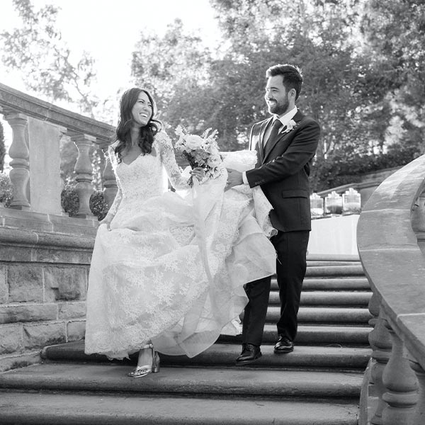 A bride and groom smile at each other as they walk up outdoor stone steps. The groom holds the bride’s dress train, and the bride carries a bouquet. Trees and sunlight are visible in the background.