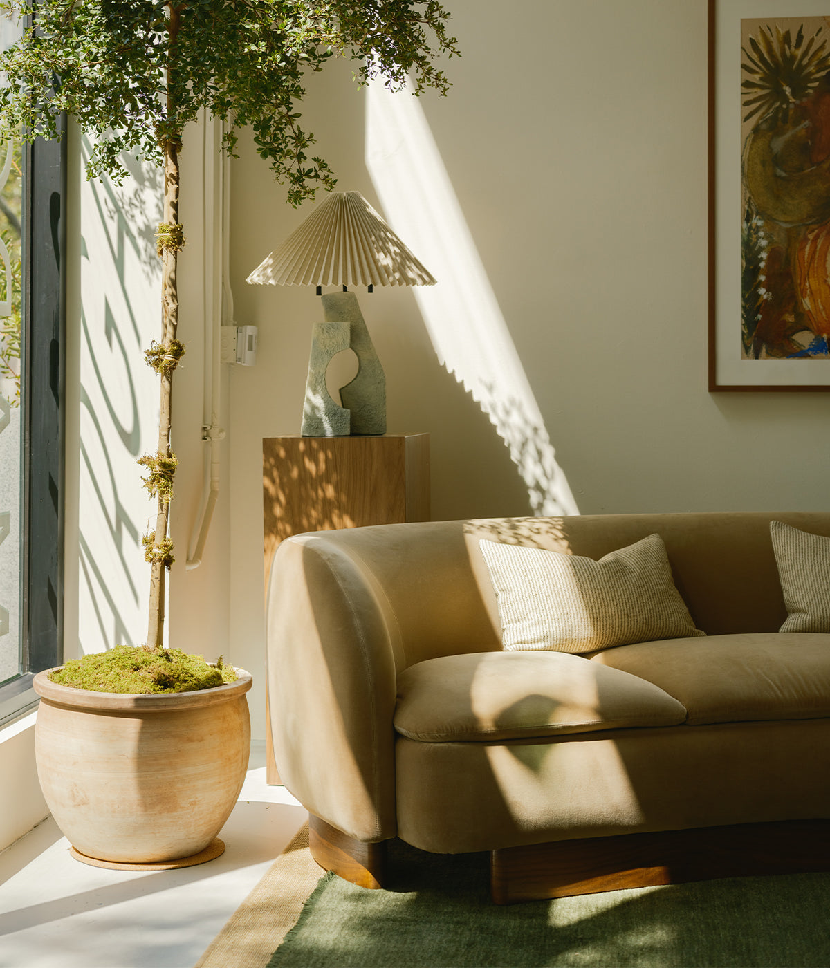 A sunlit living room with a curved beige sofa, green rug, large potted plant, sculptural table lamp on a wooden pedestal, and a colorful framed artwork on the wall. Light and shadows create a calm atmosphere.