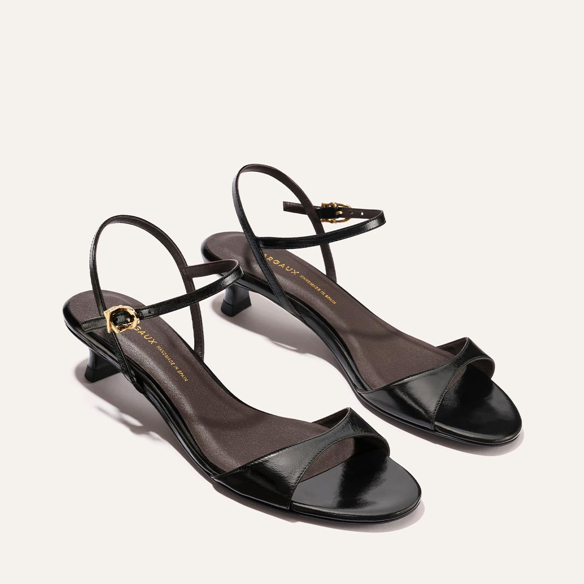 The Lotto 35 - Black Crinkle-Gloss Nappa by Margaux: black, low-heeled open-toe sandals with thin ankle straps and gold buckles, shown on a light beige background.