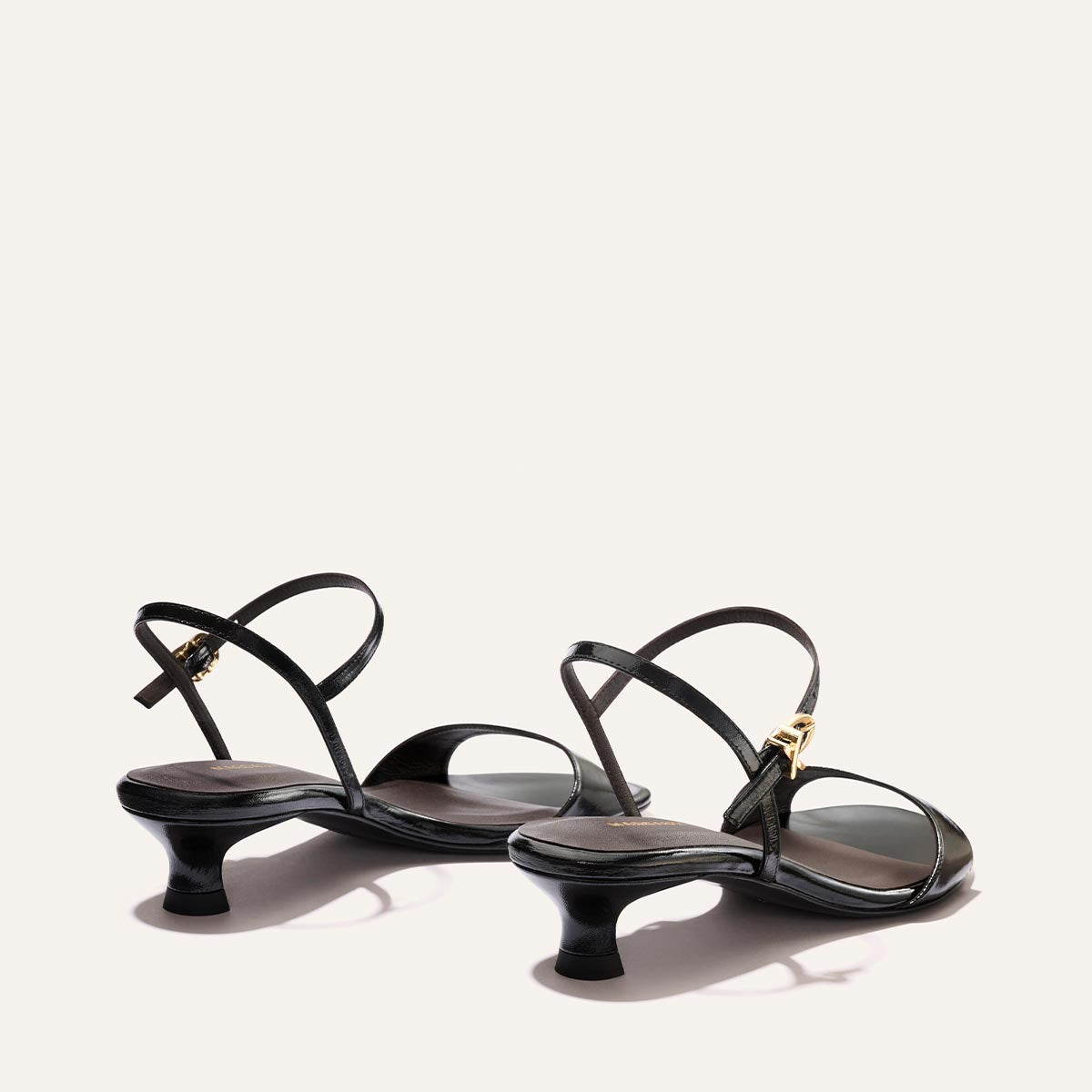 The Lotto 35 - Black Crinkle-Gloss Nappa by Margaux: black strappy heeled sandals with low, curved heels and gold buckles, shown on a plain light background from a rear-side angle.