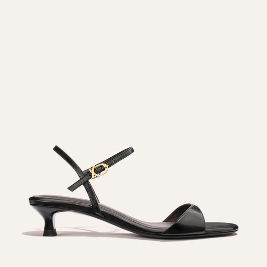 The Lotto 35 - Black Crinkle-Gloss Nappa by Margaux is a black strappy sandal with a low kitten heel, thin ankle strap with a gold buckle, and minimalist design shown against a plain light background.