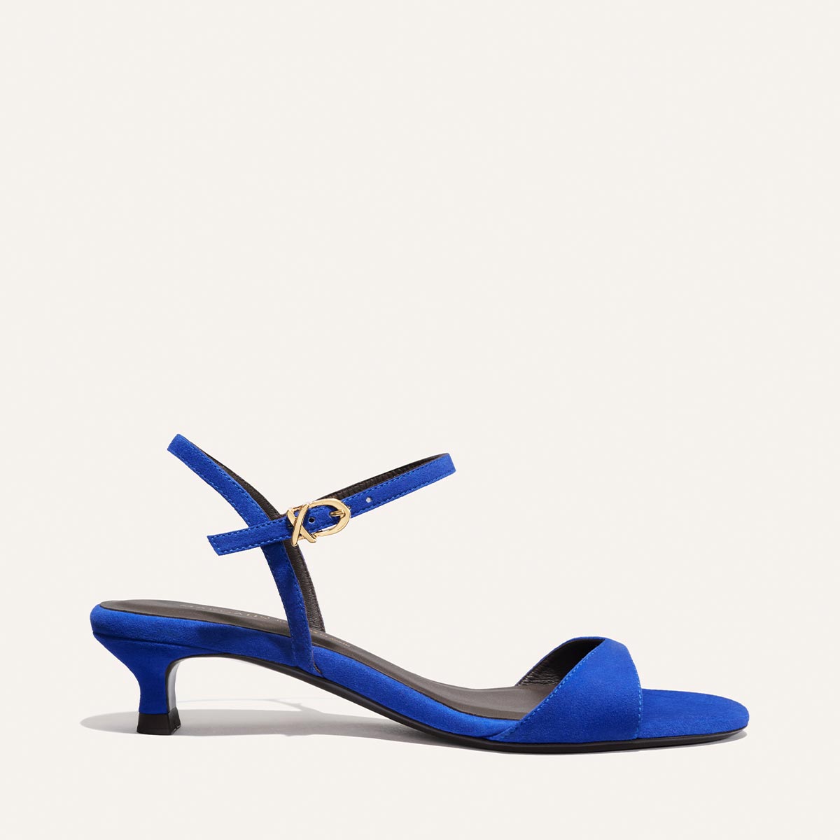 The Lotto 35 - Cobalt Suede by Margaux is a blue minimal sandal with a low kitten heel, single tapered front strap, and an ankle strap with a gold buckle, shown against a plain light background.