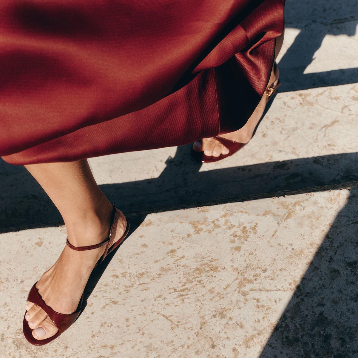 A person wearing Margaux's The Lotto 35 - Espresso Crinkle Patent sandals walks across a sunlit concrete surface in a matching satin skirt, casting distinct shadows.