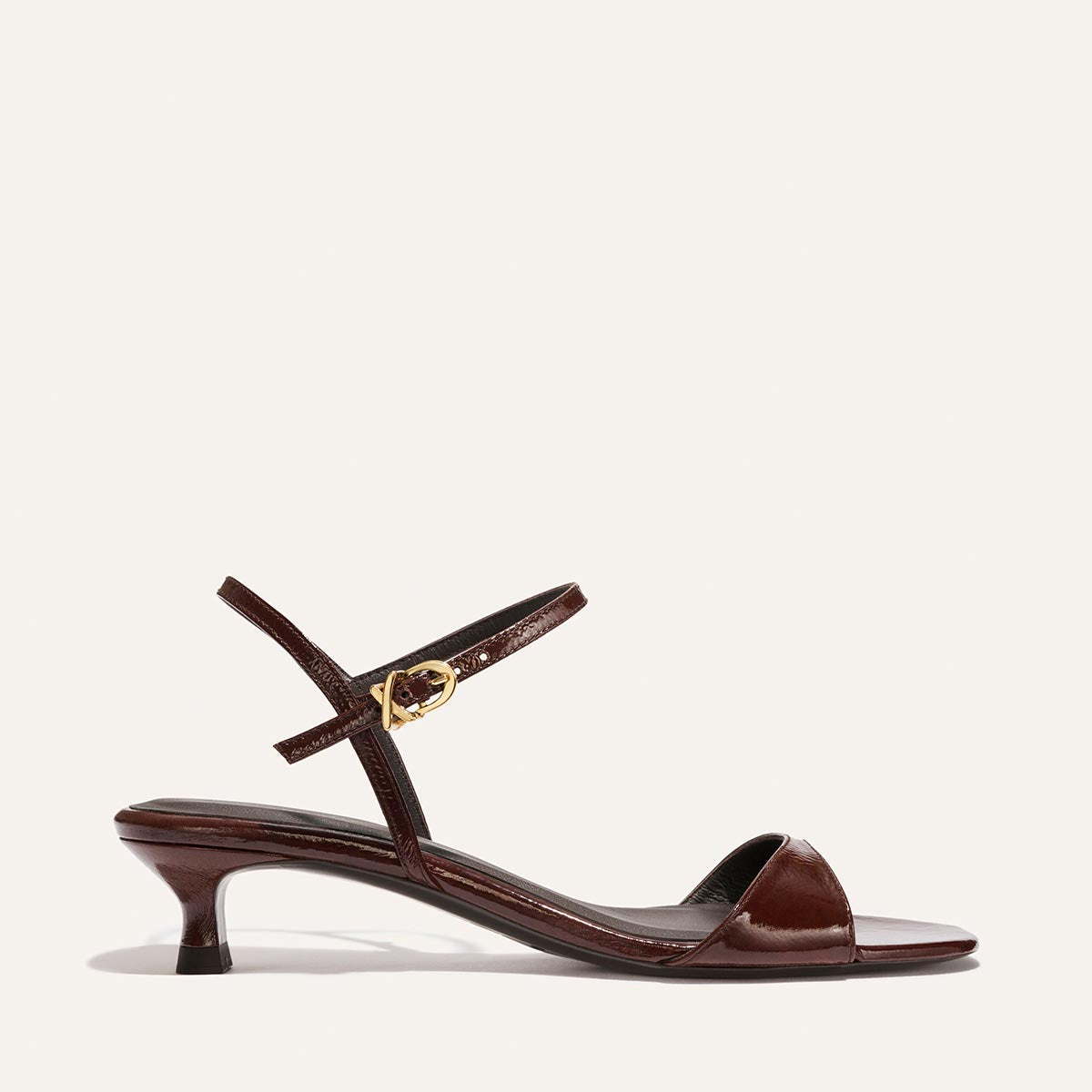 The Lotto 35 - Espresso Crinkle Patent by Margaux is a brown patent leather sandal with a low kitten heel, open toe, and slim ankle strap with a gold buckle, shown on a plain light background.