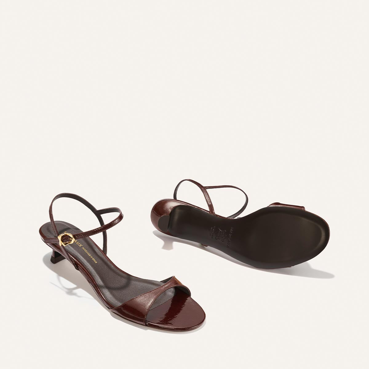 The Lotto 35 - Espresso Crinkle Patent by Margaux are heeled sandals with an open toe, thin ankle strap, and gold buckle. One shoe stands upright while the other lies on its side to display the sole and low, curved heel.