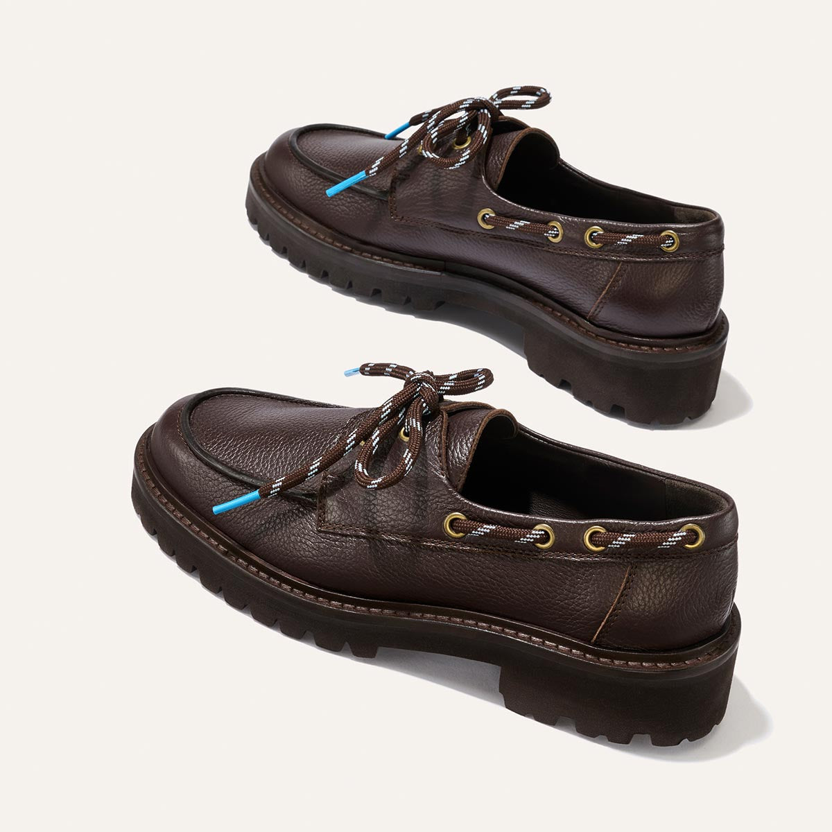The Marine Loafer - Espresso Tumbled Calf by Margaux features dark brown leather uppers, thick rugged soles, brown and white striped laces with gold eyelets, and small blue lace tips, elegantly displayed on a light background.