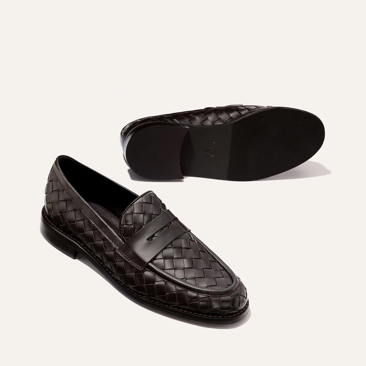A pair of The Marlowe Loafer – Brown Woven Pasado Calf by Margaux features rich dark brown woven leather uppers and sleek black soles. One loafer stands upright to showcase its modern side profile and top view, while the other lies on its side to reveal the sole—a true closet staple for any sophisticated wardrobe.