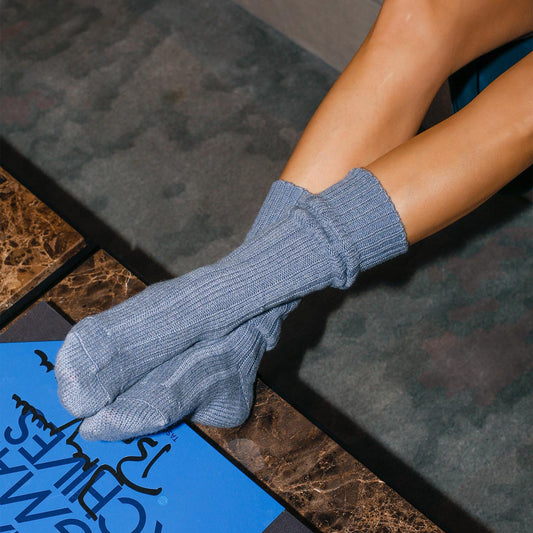 A person with bare legs lounges in Margaux’s The Cozy Sock - Blue Cashmere, feet propped on a dark marble table beside a blue magazine—ideal for cozy, luxurious relaxation.