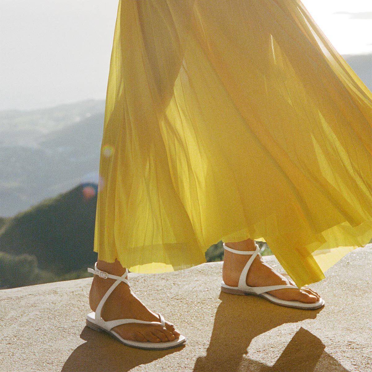 Person wearing a yellow skirt and white sandals with a scenic background