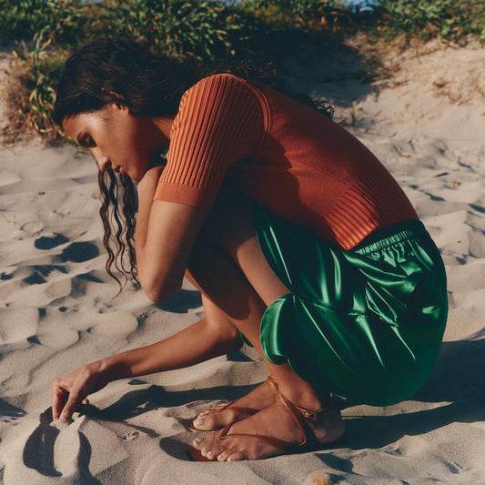 A woman with long wavy hair crouches on sandy ground, drawing in the sand amid summer greenery. She wears a short-sleeved orange top, green skirt, and Margaux’s The Palermo Sandal - Saddle Nappa with a cushioned insole.