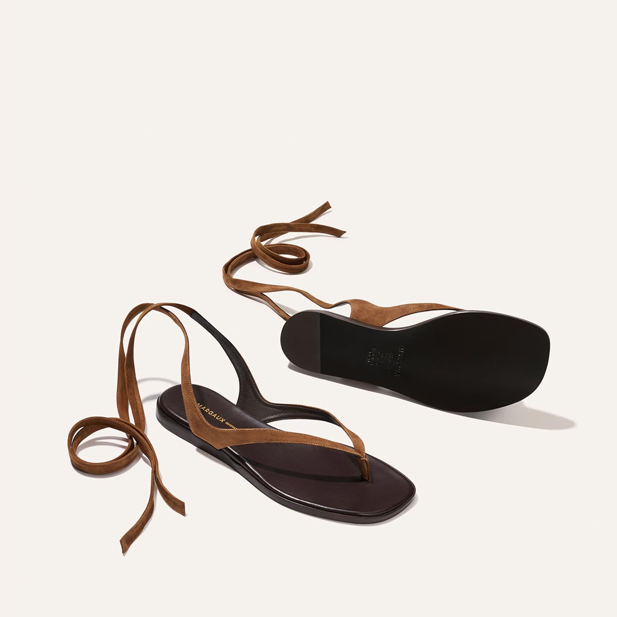 The Palermo Wrap Sandal in Nutmeg Suede by Margaux is shown with suede ankle wrap straps—one sandal upright, the other displaying the sole—all set against a light background for effortless getaway style.