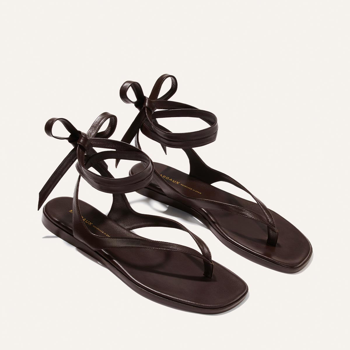 The Margaux Palermo Wrap Sandal in Espresso Nappa features thin ankle straps that tie in a bow—a chic dark brown flat perfect for summer getaways, shown on a neutral background.