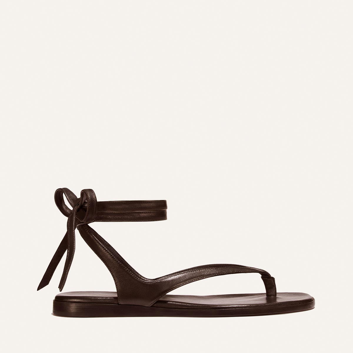The Palermo Wrap Sandal - Espresso Nappa by Margaux features minimalist black leather, a flat sole, cushioned insole, toe loop, and wrap-around ankle straps tied in a bow—perfect summer style shown on a light background.