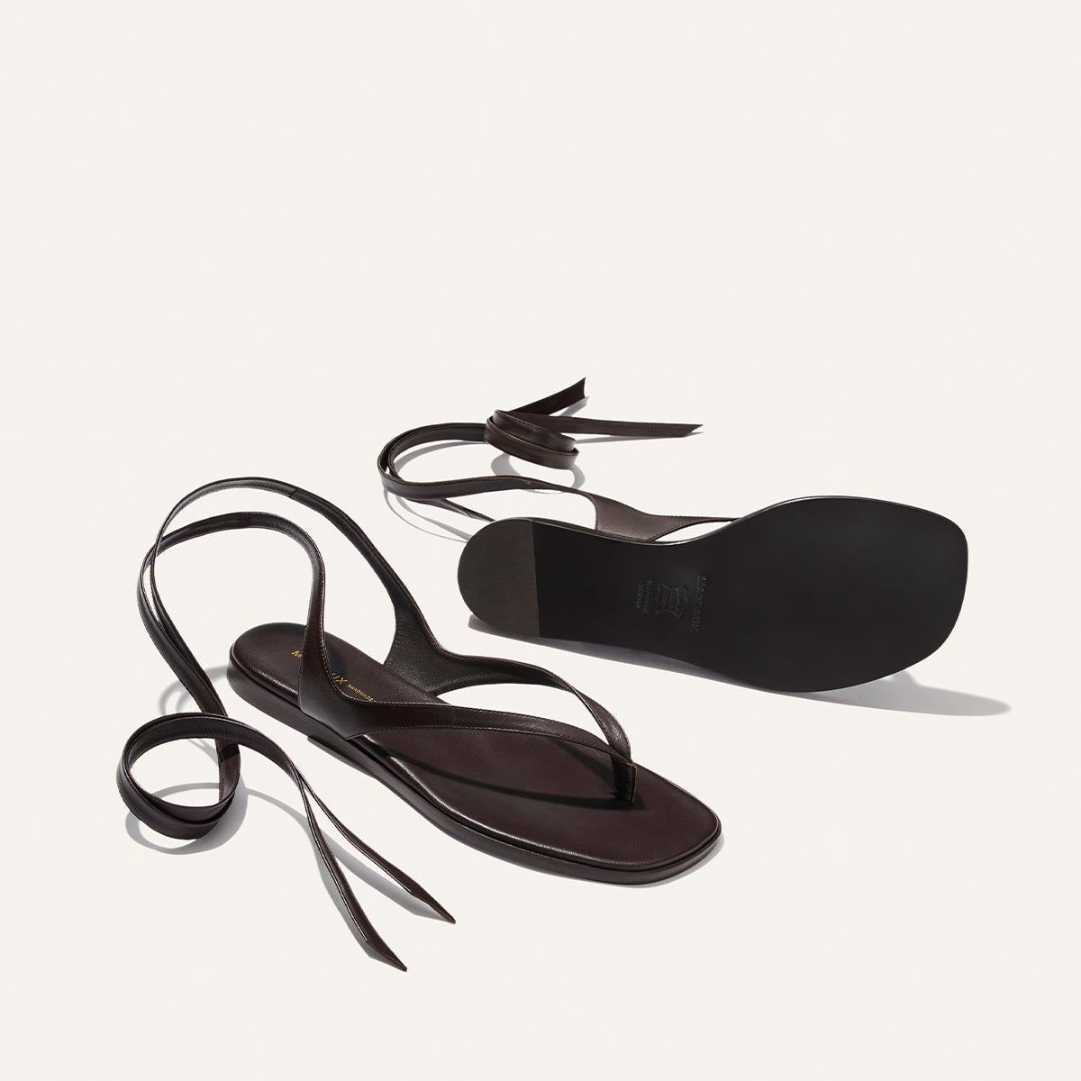 The Palermo Wrap Sandal by Margaux, in Espresso Nappa, features flat soles and long ankle wrap straps. One sandal stands upright, the other flipped to reveal its cushioned insole, all displayed against a plain white background.