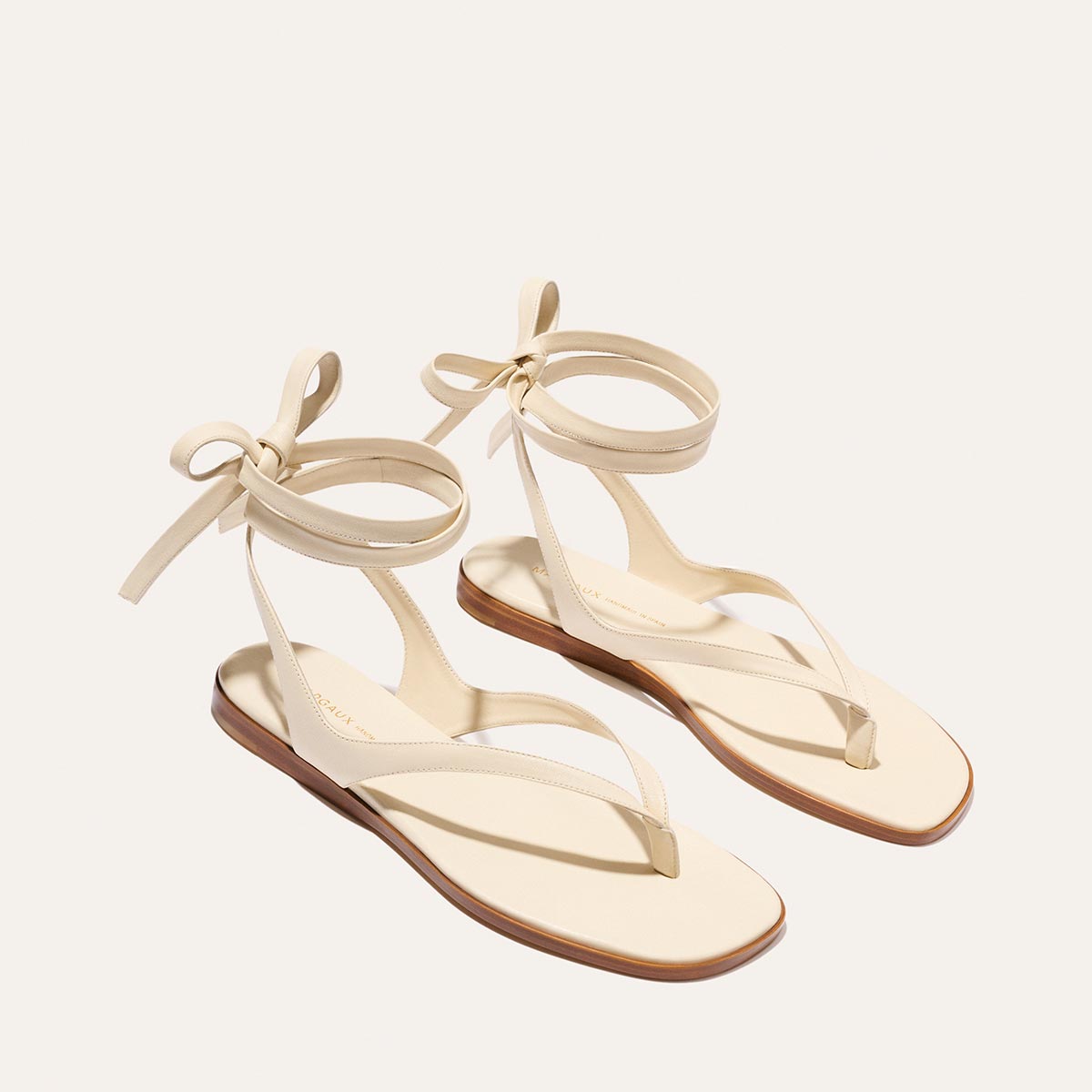 The Palermo Wrap Sandal - Ivoire Nappa by Margaux features minimal beige straps, ankle ties, and thin soles—an elegant flat sandal displayed on a light background, perfect for summer.