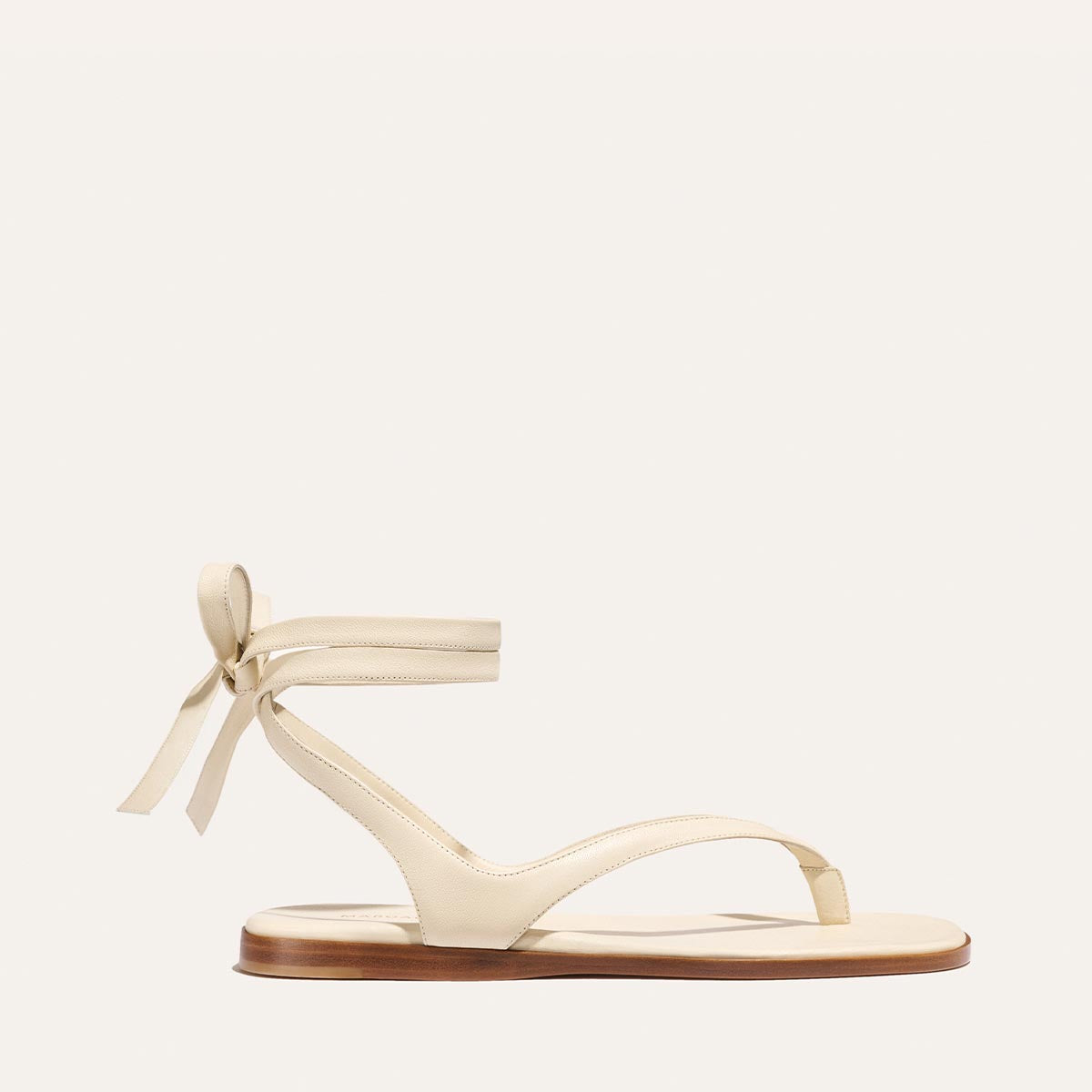 The Palermo Wrap Sandal - Ivoire Nappa by Margaux features cream-colored thong straps, elegant side-knotted ankle ties, and a flat brown sole. Shown on a plain off-white background.