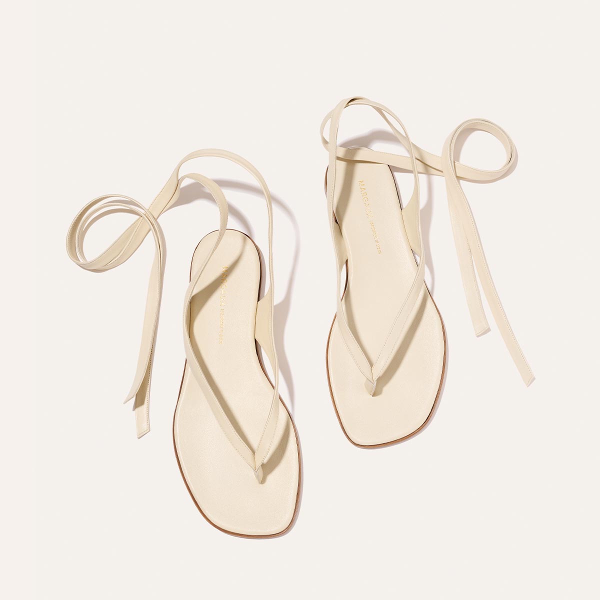 The Palermo Wrap Sandal - Ivoire Nappa by Margaux is a cream minimalist summer sandal with long ankle ties, square soles, and thin thong-style uppers, shown on a light background for an effortlessly chic look.