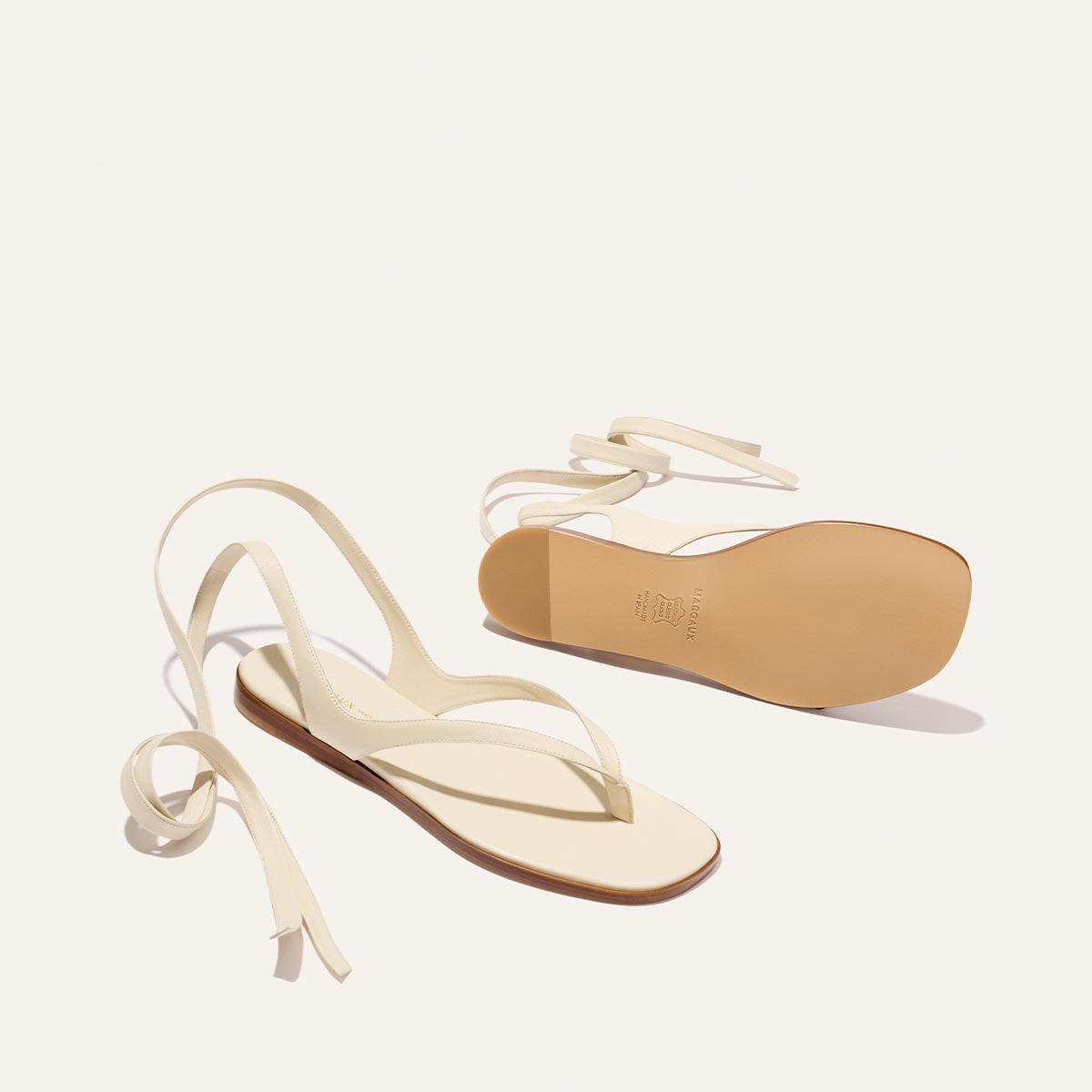 The Palermo Wrap Sandal - Ivoire Nappa by Margaux features minimalistic straps and thin ankle ties. One sandal is displayed top-down, the other sole-up, both set against a plain off-white background.