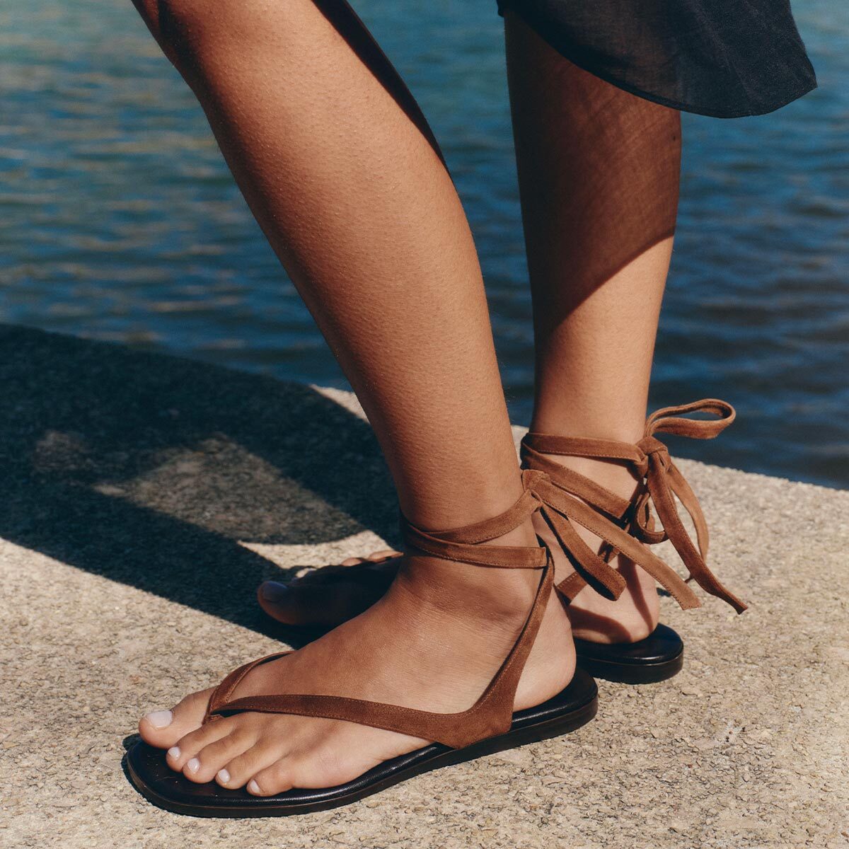 A person wearing Margaux's Palermo Wrap Sandal in Nutmeg Suede stands on a concrete surface by the water, with part of a dark skirt visible. Sunlight creates shadows across their legs.