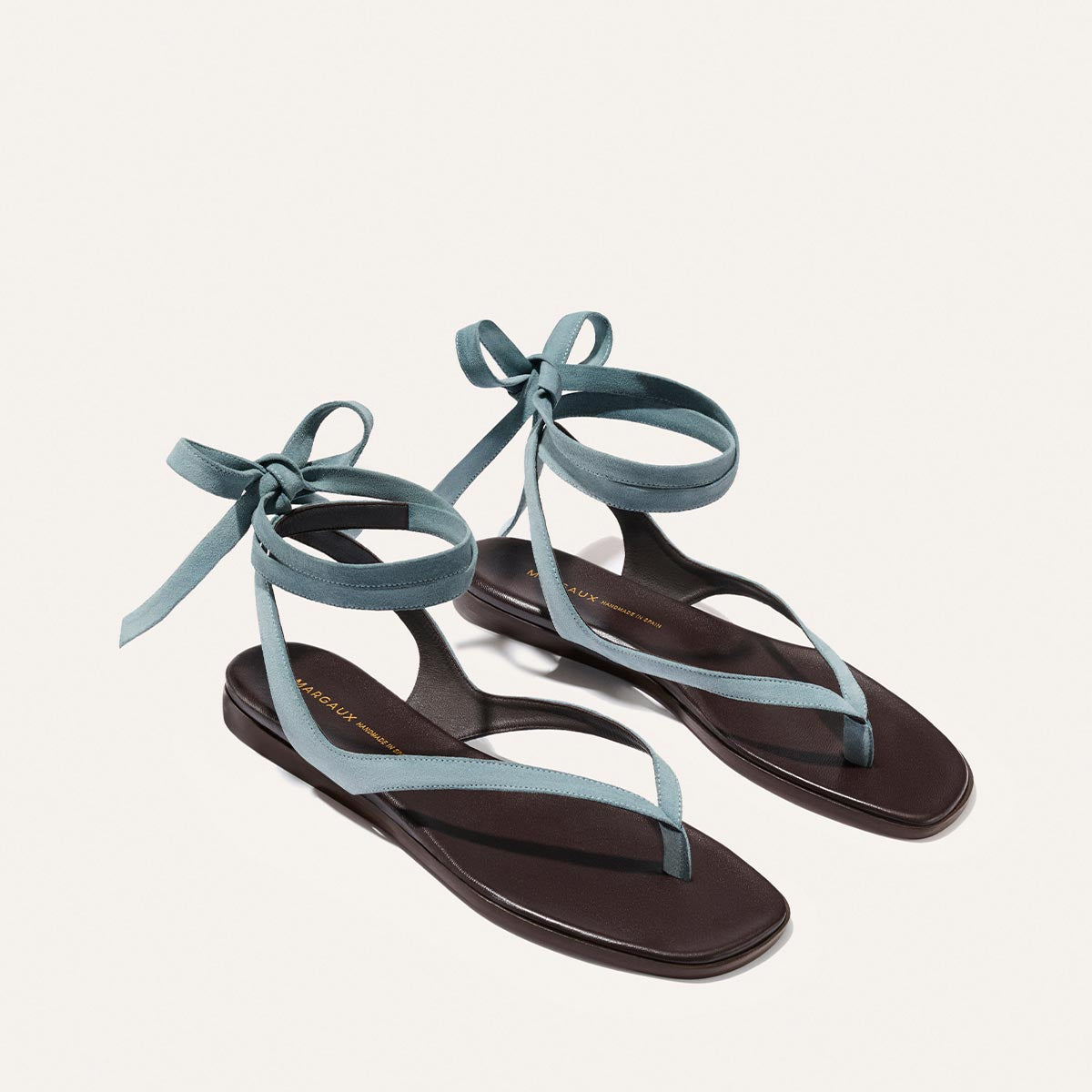 The Margaux Palermo Wrap Sandal in Ocean Suede features flat, dark brown soles with thin, blue-green crisscross straps and ankle ties, shown on a light background.