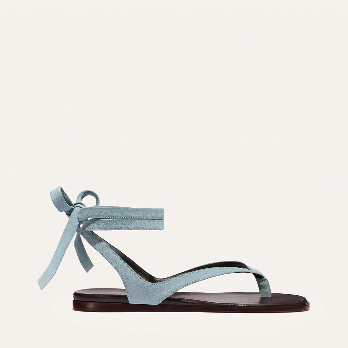 The Margaux Palermo Wrap Sandal in Ocean Suede features a light blue upper, flat dark sole, open toe, and ankle wrap with bow, displayed against a plain off-white background.