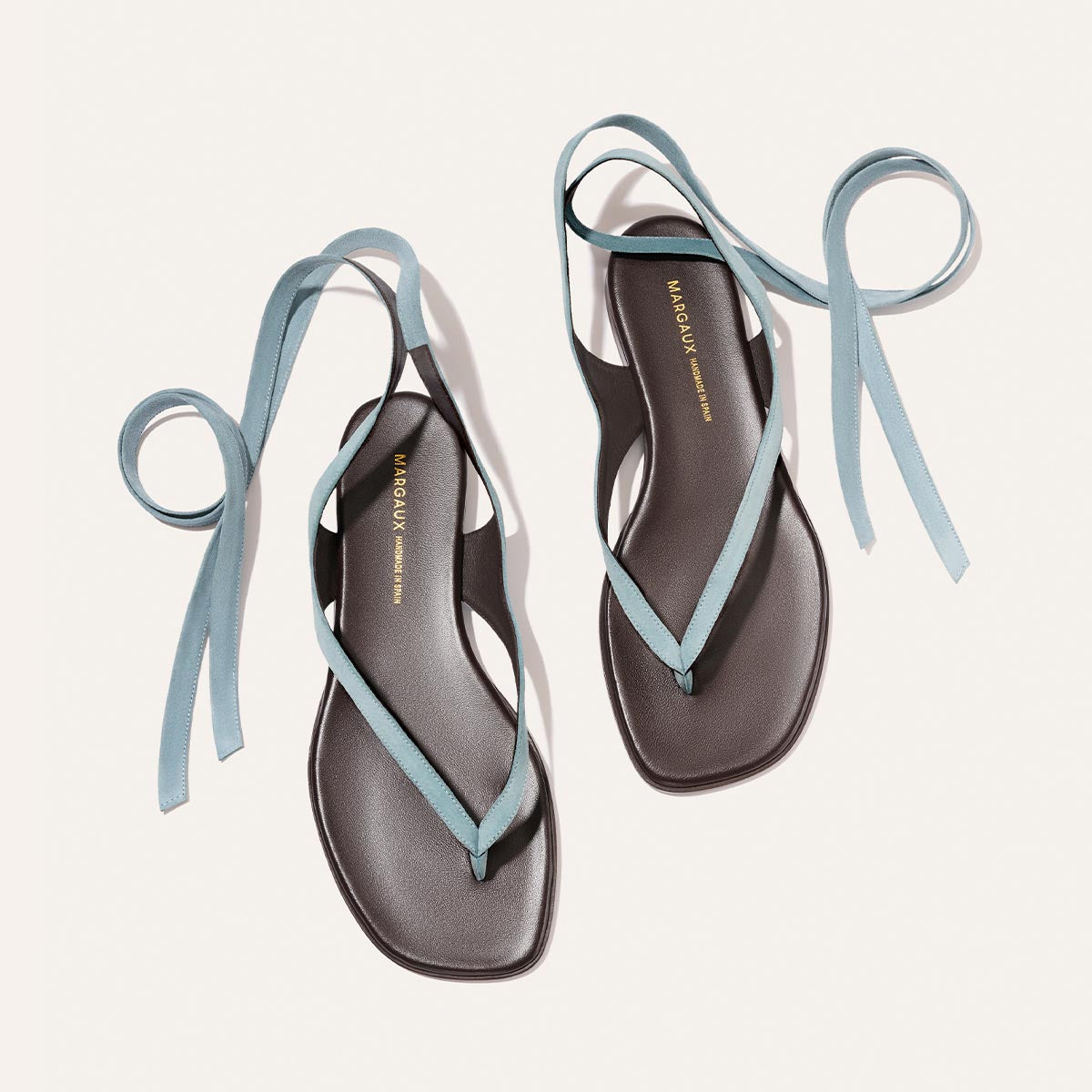 A pair of The Palermo Wrap Sandal in Ocean Suede by Margaux, featuring black square toes and light blue ankle wrap straps, is displayed on a white background. The Margaux brand name appears on each sandal’s insole in gold lettering.