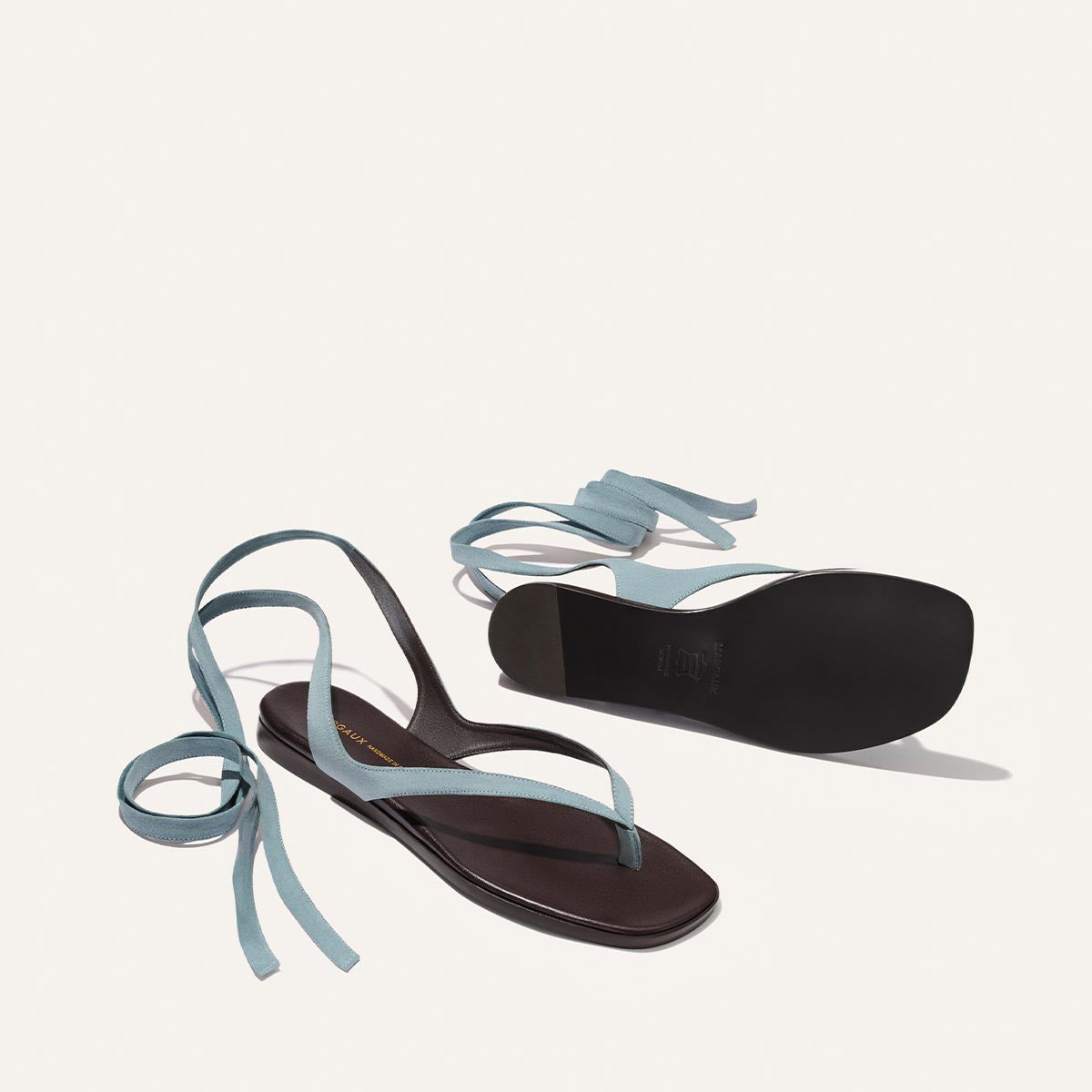 The Margaux Palermo Wrap Sandal in Ocean Suede features black sandals with square toes and thin blue ankle wrap straps, shown on a white background—one viewed from the top, the other revealing the sole.