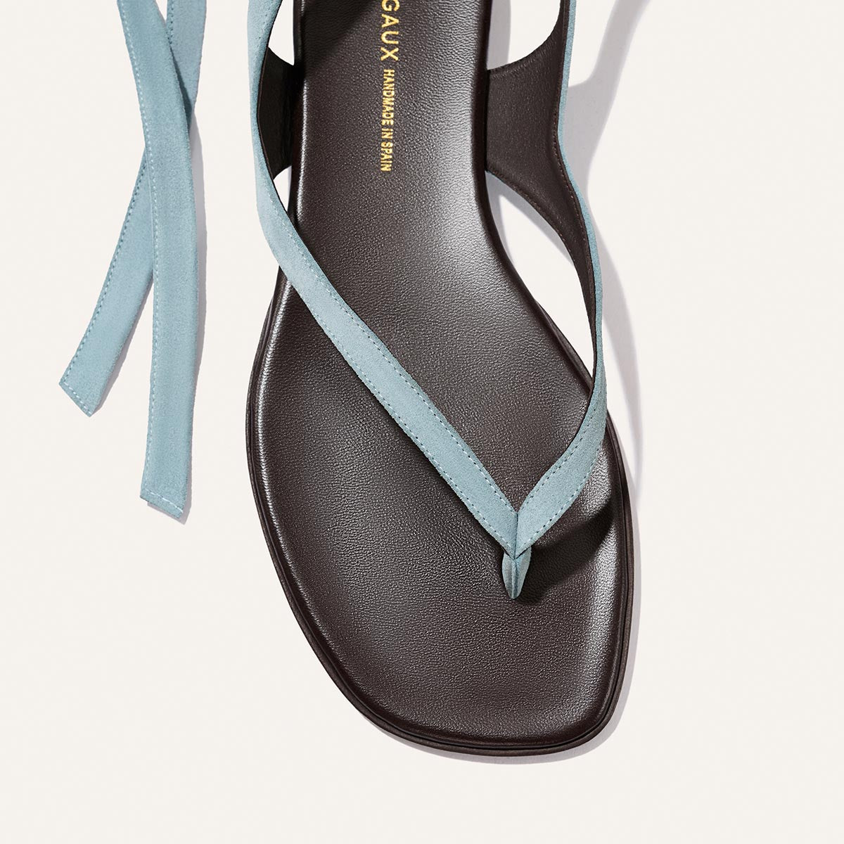 A close-up of Margaux's The Palermo Wrap Sandal in Ocean Suede, featuring a black square toe and light blue suede straps that loop between the toes and wrap around the ankle.