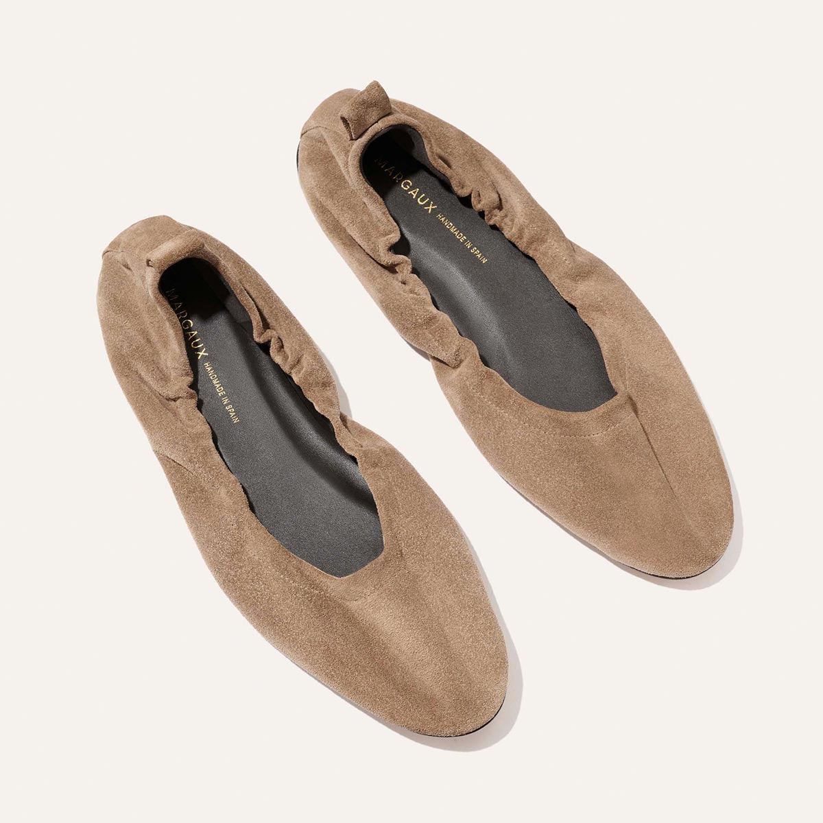 The Pascale - Cappuccino Suede ballet flats by Margaux feature taupe suede, an elasticated back, and black insoles, shown on a light background.