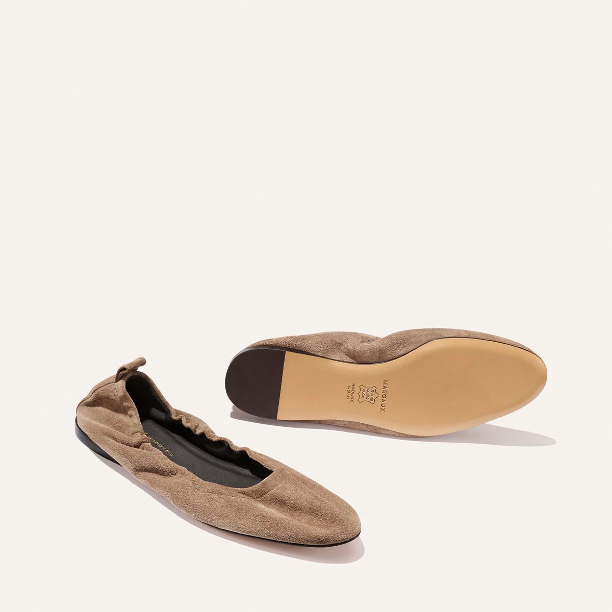 The Margaux Pascale - Cappuccino Suede features light brown suede ballet flats with ruched details, an elasticated back, and a small bow at the heel; one shoe stands upright while the other displays the sole.