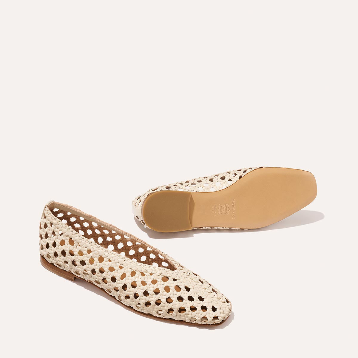 The Woven Paz SS25 - Ecru Leather by Margaux are cream-colored, handwoven flats featuring pointed toes and cut-out details. One shoe is upright, revealing the top and side; the other lies on its side to display the sole.