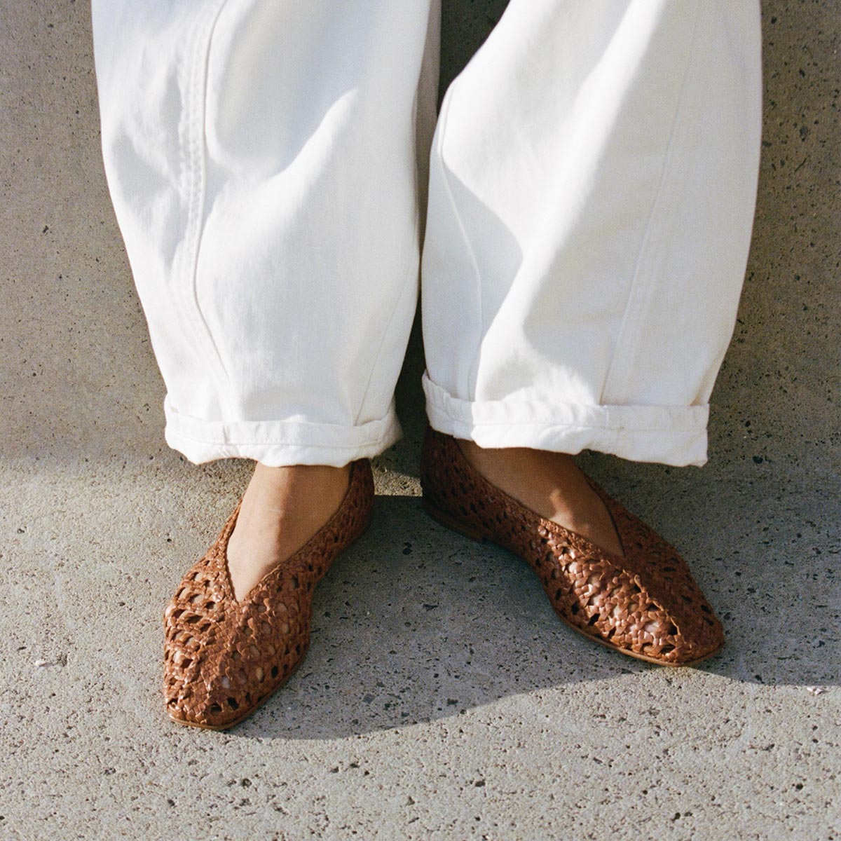 A person stands on a concrete surface wearing luxuriously soft, loose white pants with a higher rise and Margaux’s The Woven Paz flats in Saddle Woven Leather.