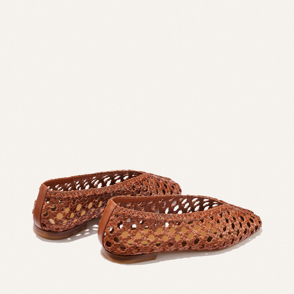 The Woven Paz - Saddle Woven Leather by Margaux are brown, handwoven leather flats with a perforated lattice design, closed toes, and low heels. Shown against an off-white background, they are simple and lightweight.